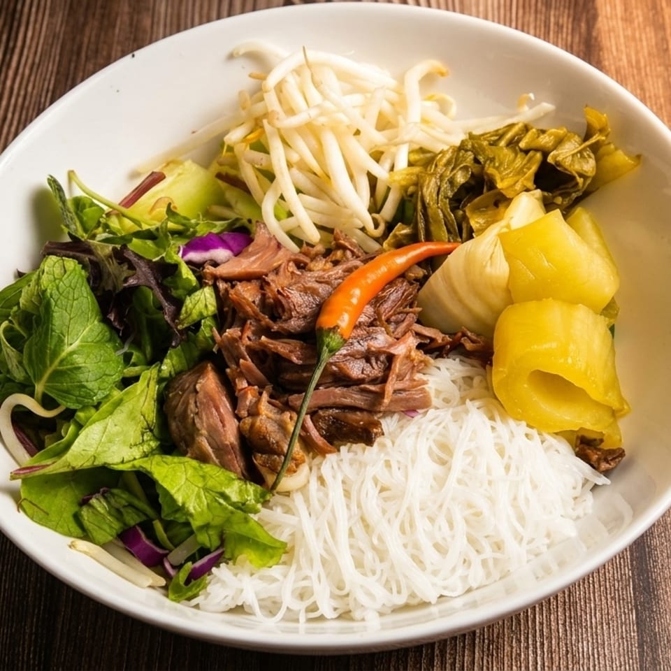 A white bowl filled with vermicelli noodles, shredded meat, bean sprouts, and greens on a wooden table.
